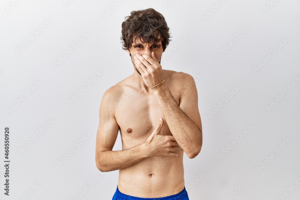 Young hispanic man standing shirtless over isolated, background smelling something stinky and disgusting, intolerable smell, holding breath with fingers on nose. bad smell
