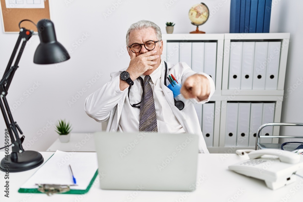 Senior caucasian man wearing doctor uniform and stethoscope at the clinic laughing at you, pointing finger to the camera with hand over mouth, shame expression Senior caucasian man wearing doctor uniform and stethoscope at the clinic laughing at you, pointing finger to the camera with hand over mouth, shame expression