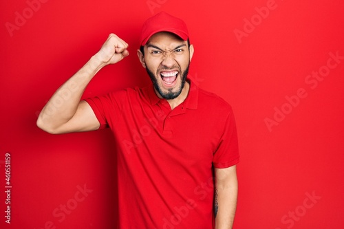 Hispanic man with beard wearing delivery uniform and cap angry and mad raising fist frustrated and furious while shouting with anger. rage and aggressive concept.