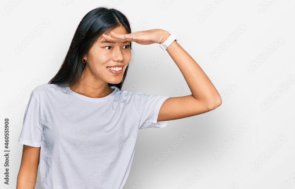 Beautiful young asian woman wearing casual white t shirt very happy and smiling looking far away with hand over head. searching concept. Beautiful young asian woman wearing casual white t shirt very happy and smiling looking far away with hand over head. searching concept.