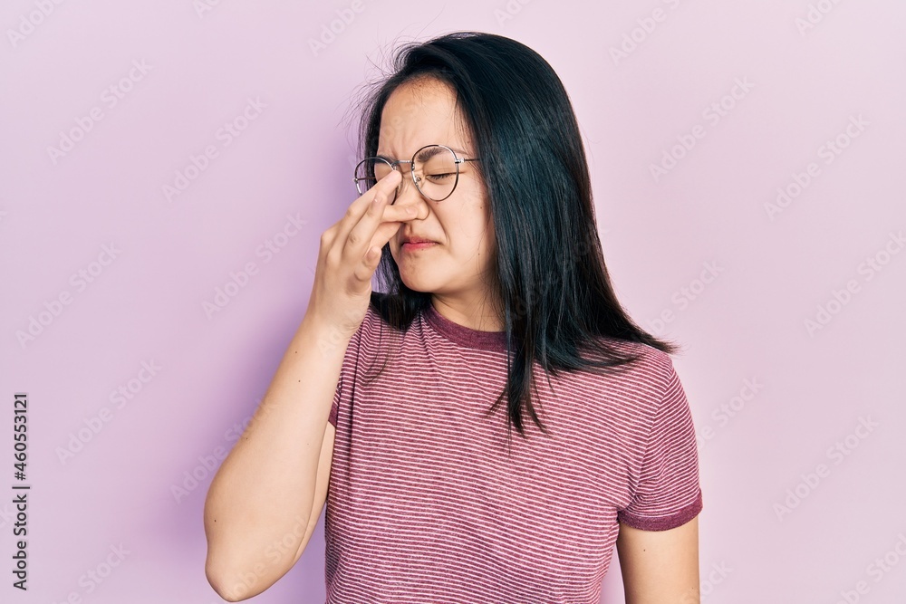 Young chinese girl wearing casual clothes and glasses smelling something stinky and disgusting, intolerable smell, holding breath with fingers on nose. bad smell Young chinese girl wearing casual clothes and glasses smelling something stinky and disgusting, intolerable smell, holding breath with fingers on nose. bad smell