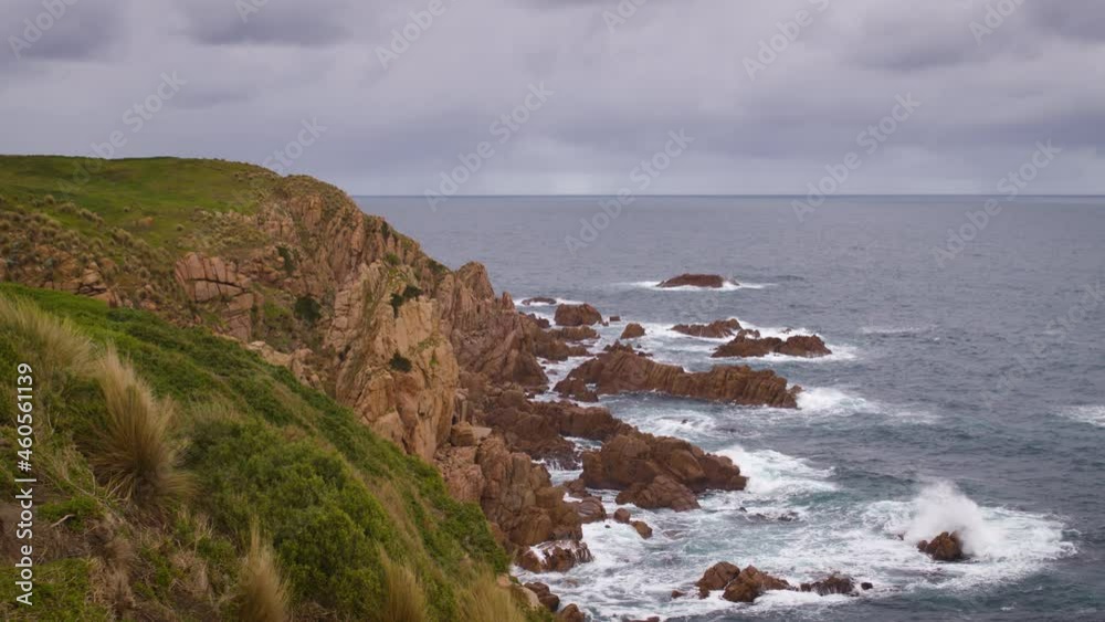 Waves breaking against red rocky coastline with green hills, a scenic view of the vast ocean and storm clouds in the background. Footage captured at the pinnacles on Phillip Island in Australia.
