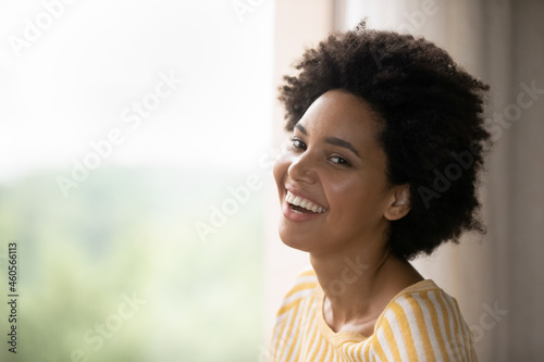 Head shot attractive young african american woman laughing standing near window, daydreaming visualizing future, planning weekend, imagining opportunity at home, copy space for advertisement.