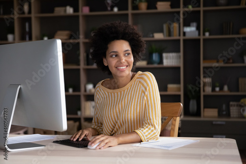 Thoughtful smiling young african american business lady working on computer in modern home office, looking in distance considering project problem solution or creating new ideas, feeling motivated.