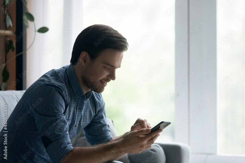 Side view smiling man using smartphone, sitting on cozy couch at home, happy young male looking at phone screen, chatting online, typing writing message in social network, shopping, surfing internet Side view smiling man using smartphone, sitting on cozy couch at home, happy young male looking at phone screen, chatting online, typing writing message in social network, shopping, surfing internet