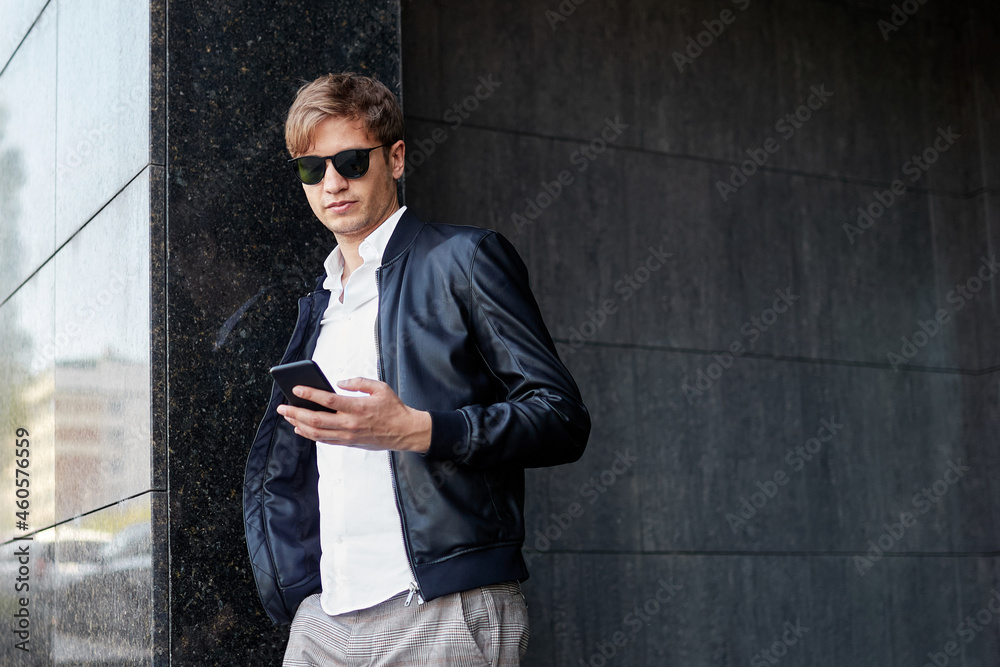 Confident young handsome business man wearing sunglasses, posing outside near building, read a message in his phone. Confident young handsome business man wearing sunglasses, posing outside near building, read a message in his phone.
