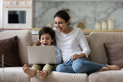 Joyful young indian woman cuddling laughing small cute kid son, watching together funny comedian movie or cartoons online on laptop, relaxing on couch, tech addiction concept.