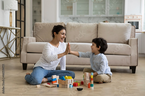 Fotografie Happy millennial asian ethnic mother or nanny giving high five to little child boy, playing with toy bricks in modern living room, cheerful indian family entertaining, sitting on wooden floor at home