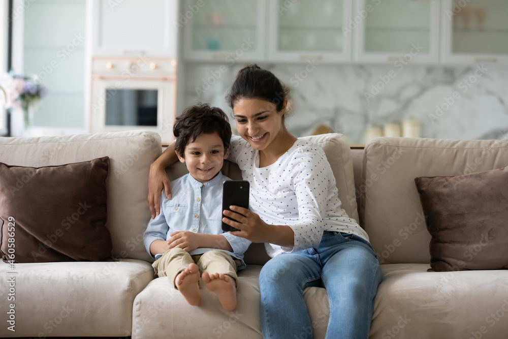 Smiling bonding young indian woman showing funny mobile applications to adorable little child son, recording streaming online stories for social network, posing for selfie photo, or playing games.