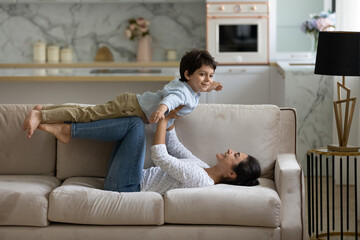Happy young indian mother lifting in air on straight arms joyful cute small child son, lying on cozy couch. Joyful asian family practicing balance doing yoga exercises, having fun playing at home.