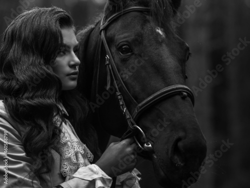 A young beautiful brunette rider next to a black mare in full ammunition,