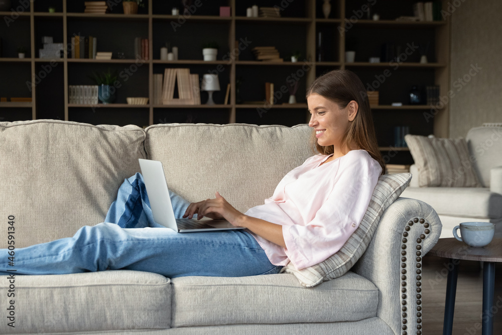 Happy millennial girl with laptop relaxing on couch in living room, typing, smiling, chatting online, browsing internet. Blogger, marketer writing article. Student studying at home, watching webinar Happy millennial girl with laptop relaxing on couch in living room, typing, smiling, chatting online, browsing internet. Blogger, marketer writing article. Student studying at home, watching webinar