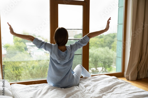 Young woman waking up in bed with soft comfortable mattress, stretching arms after good sleeping, bedtime recreation. Tourist woman enjoying vacation, looking out of window from hotel room. Back view