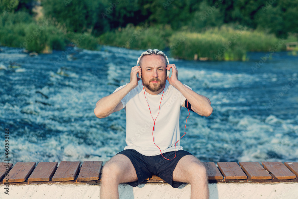 A man with a beard and a braid haircut listens to music with headphones on the river bank