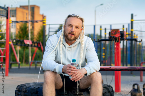 A man with an interesting braid hairstyle drinks water from a bottle after a workout on an outdoor fitness site