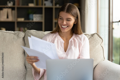 Happy young student girl receiving admission letter from business school, college, sitting on couch, holding laptop, reading paper document, smiling, laughing, feeling joy, celebrating success photo