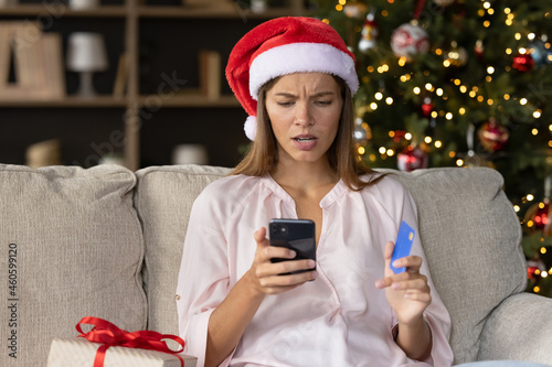 Worried concerned girl in Christmas Santa hat having problems with payment by credit card online for New Year purchases, looking at smartphone screen with puzzled face. Scam, fraud concept