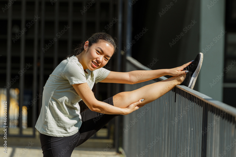 Positive young Asian woman in stylish tracksuit does leg stretching exercise leaning on bridge railing in sunny city street