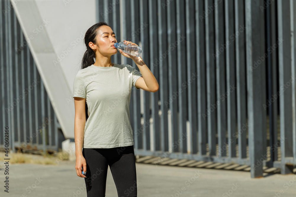 Athletic Asian lady with ponytail drinks water from plastic bottle resting after training on sunny city street Athletic Asian lady with ponytail drinks water from plastic bottle resting after training on sunny city street