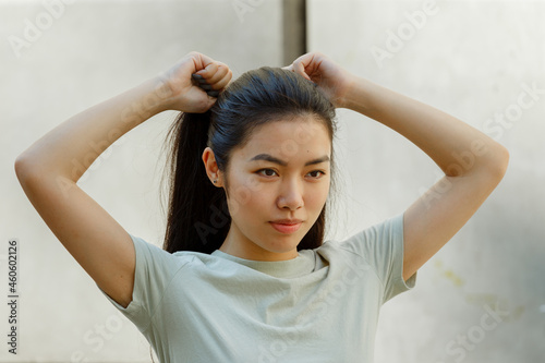 Pretty Asian young woman in t-shirt adjusts ponytail made of long dark hair standing near white wall on sunny day close view