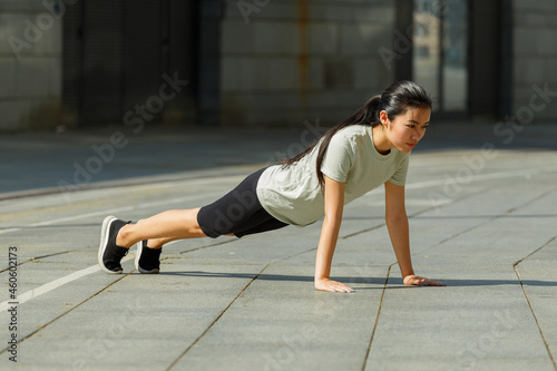 Slim Asian sportswoman in tracksuit stands in high plank posture on pavement near handrail at outdoor training on city street
