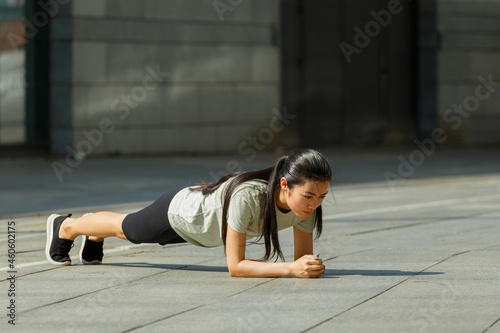 Slim Asian sportswoman in tracksuit stands in low plank posture on pavement near handrail at outdoor training on city street
