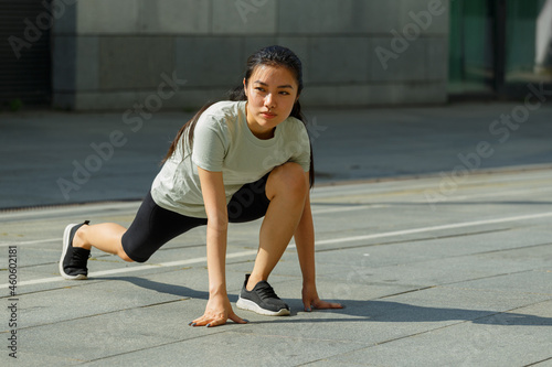 Pretty young Asian sportswoman in tracksuit does forward dynamic lunges during outdoor training on sunny city square closeup