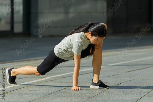 Pretty young Asian sportswoman in tracksuit does forward dynamic lunges during outdoor training on sunny city square closeup