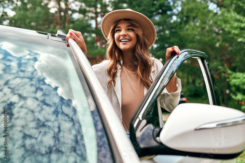A cute woman in a hat stands near the car on the background of the forest. Purchase, car rental. Rest in nature, weekend.