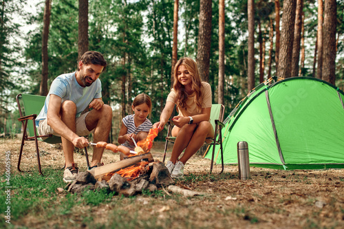 Happy family with a child on a picnic sit by the fire near the tent and grill a barbecue in a pine forest. Camping, recreation, hiking.