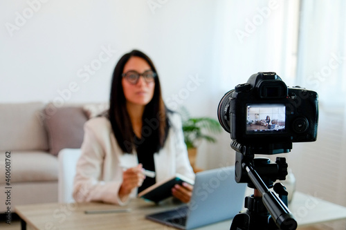 Young smart looking woman wearing glasses recording a video lesson on a camera. Portrait of female teacher by the desk with a textbook explaining a subject. Close up, copy space, brick wall background