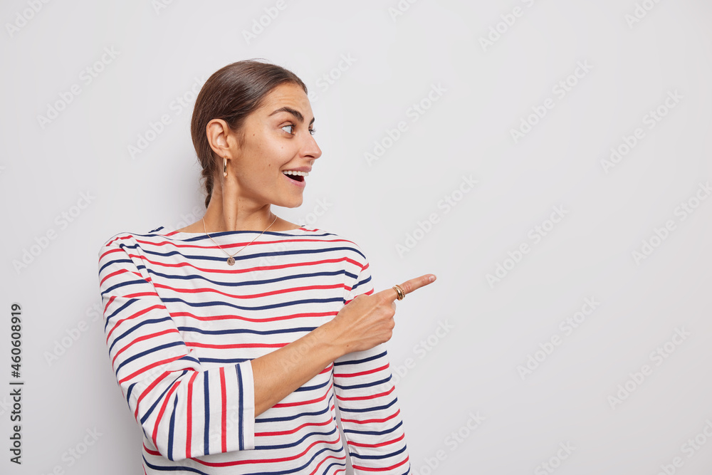 Horizontal shot of cheerful dark haired woman dressed in casual striped jumper points on right at blank copy space shows advertisement isolated over white background reads logo sign shows sale Horizontal shot of cheerful dark haired woman dressed in casual striped jumper points on right at blank copy space shows advertisement isolated over white background reads logo sign shows sale