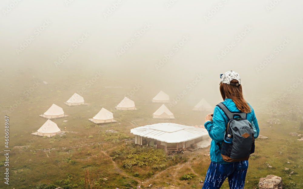 Obraz premium rear view of young woman in cap and blue sportswear with backpack hiker standing in green mountain valley and looking at tent camping in fog, hiking
