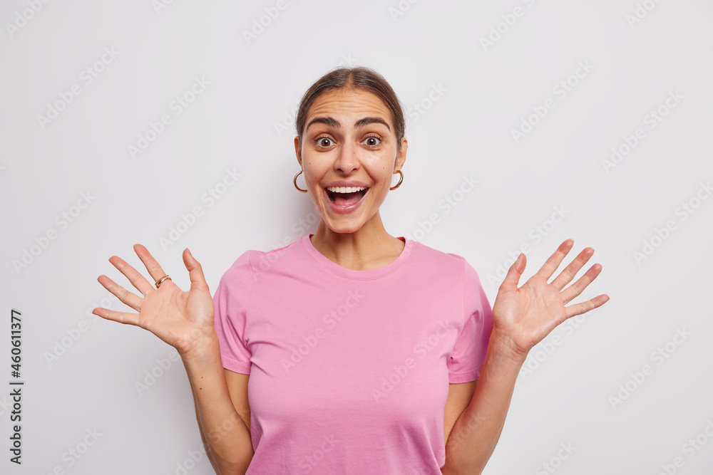 Happy cheerful woman raises palms feels glad smiles cheerfully reacts on amazing news dressed in casual pink t shirt isolated over white background. People human reactions and feelings concept Happy cheerful woman raises palms feels glad smiles cheerfully reacts on amazing news dressed in casual pink t shirt isolated over white background. People human reactions and feelings concept