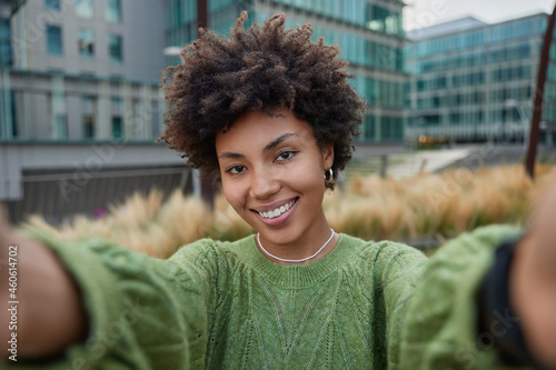 Positive Afro American woman keeps arms outstretched makes selfie photo during leisure time photographs herself during free day outdoors wears green sweater smiles broadly looks cheerfully at camera