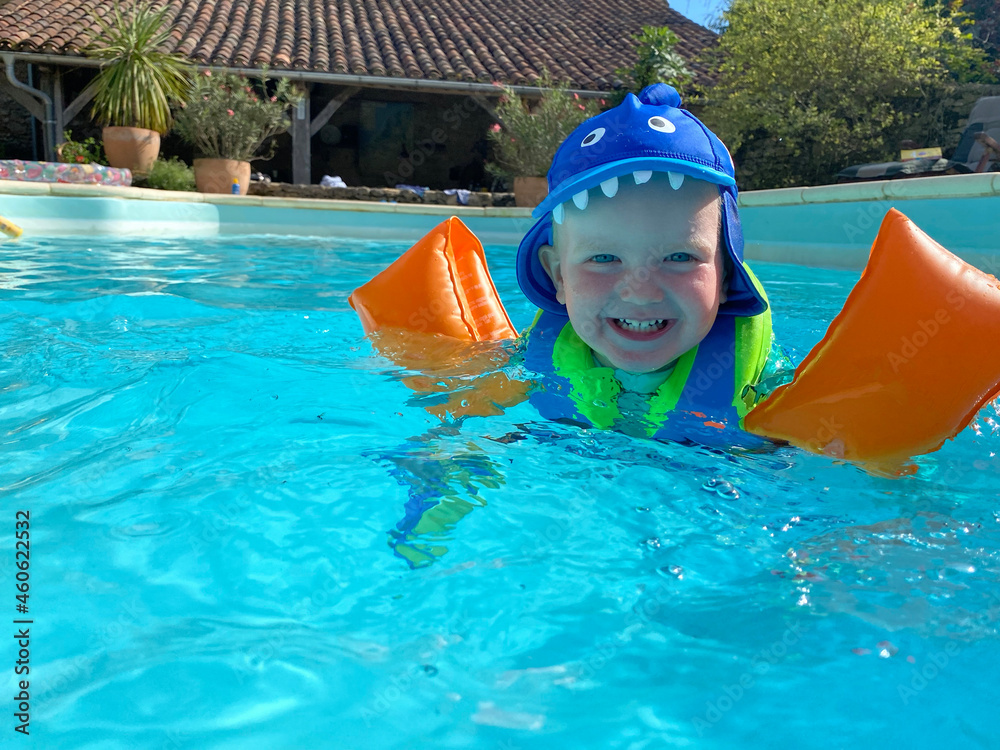 A two year old boy wearing arm bands splashes and plays in a swimming pool during summer