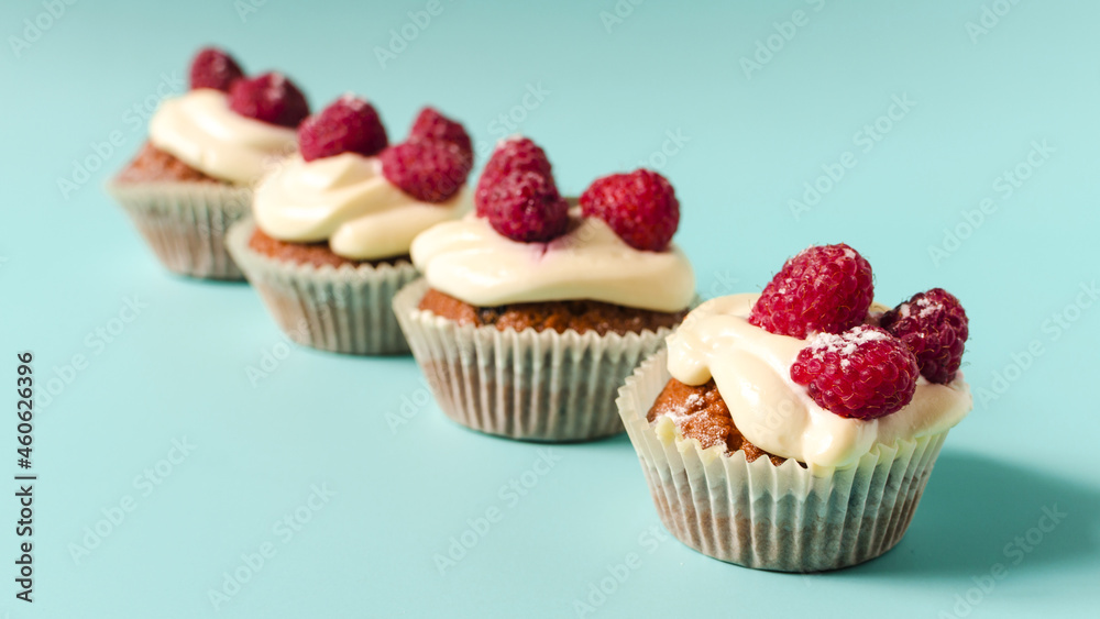 Cupcakes with white cream and raspberries lined up on a blue background sharpness in the foreground. Sweet homemade cupcakes on a colored background close-up.