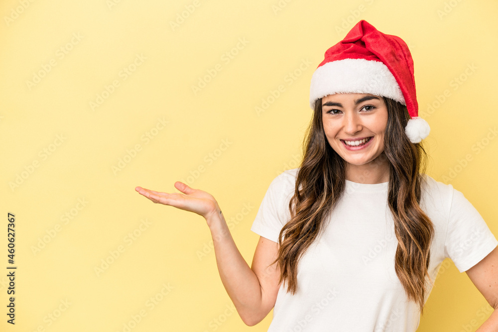 Young caucasian woman celebrating Christmas isolated on yellow background showing a copy space on a palm and holding another hand on waist. Young caucasian woman celebrating Christmas isolated on yellow background showing a copy space on a palm and holding another hand on waist.