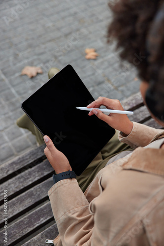Unrecognizable skilled woman designer works on digital tablet downloads application draws with stylus poses outdoor on wooden bench. Blank empty screen for your information or advertising text.