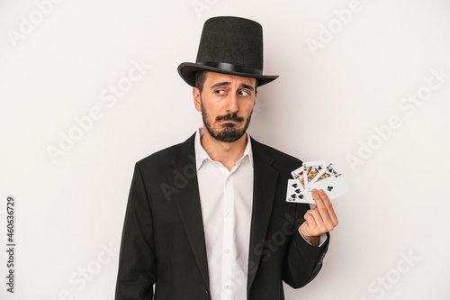 Young magician man holding a magic card isolated on white background confused, feels doubtful and unsure.