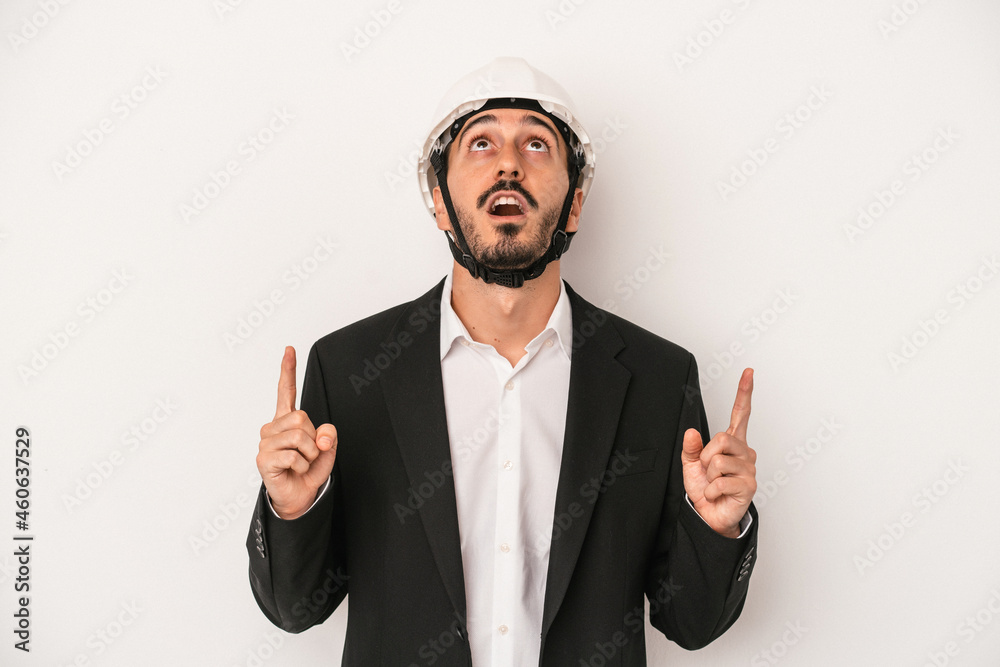 Young architect man wearing a construction helmet isolated on white background pointing upside with opened mouth. Young architect man wearing a construction helmet isolated on white background pointing upside with opened mouth.