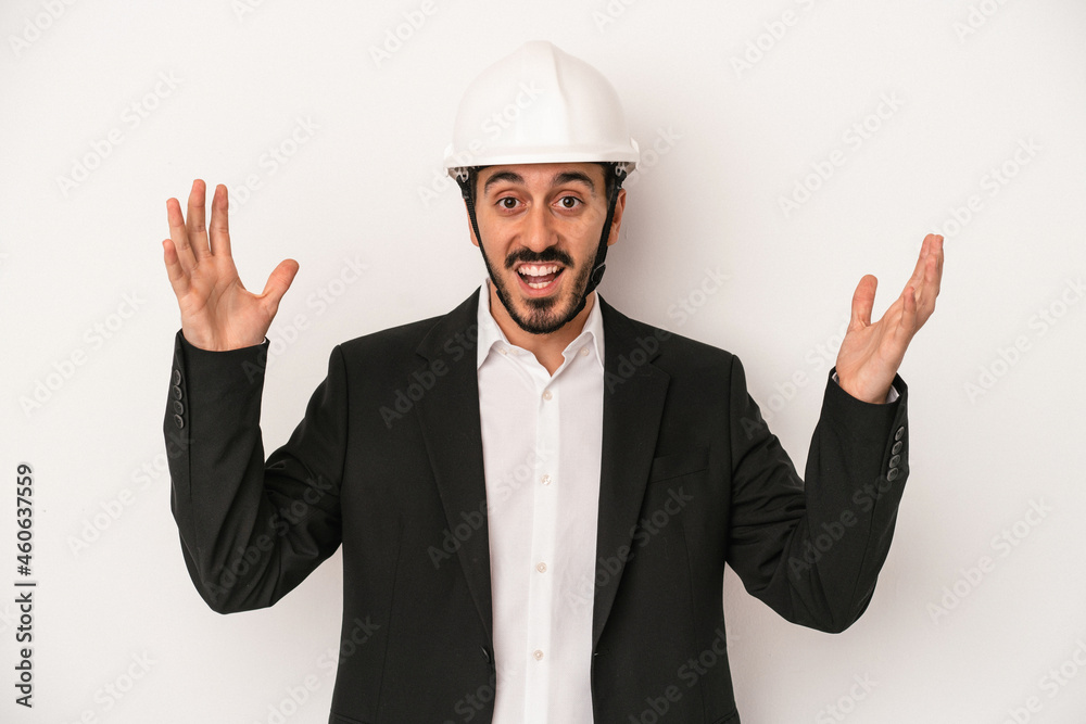 Young architect man wearing a construction helmet isolated on white background receiving a pleasant surprise, excited and raising hands.