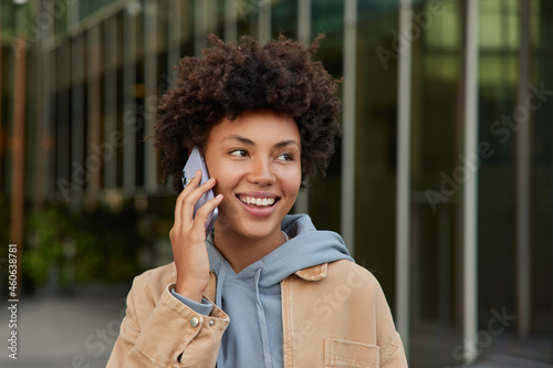Pretty positive Afro American woman laughs while calls on smartphone talks in roaming has curly hair dressed in casual clothes poses outside over blurred background makes international communication