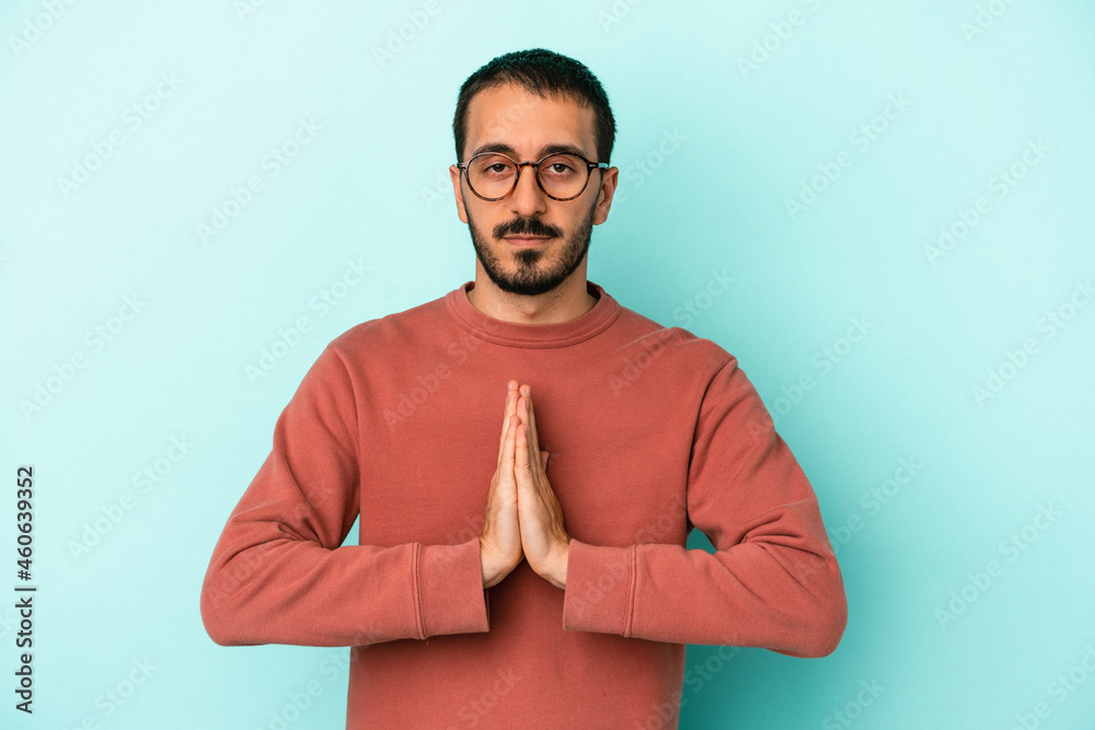 Young caucasian man isolated on blue background praying, showing devotion, religious person looking for divine inspiration.