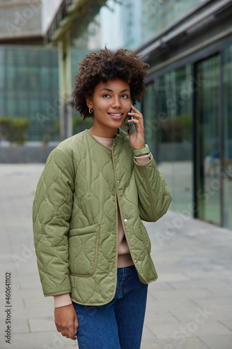 Vertical image of pleased happy Afro American woman enjoys positive cell phone calling smiles pleasantly looks into distance wears casual outfit has walk in urban setting satisfied with cheap tariffs