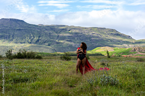 African American woman with afro hair is wearing a fashionable cosplay red dress while walking along the green landscape in Iceland. the independent woman is bravely taking on challenges