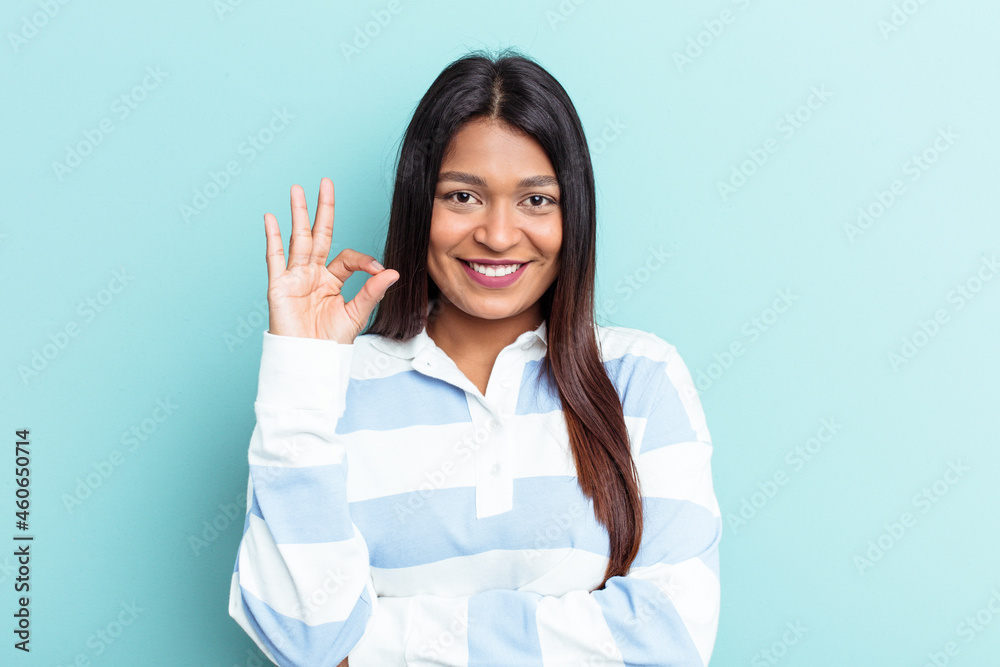 Young Venezuelan woman isolated on blue background winks an eye and holds an okay gesture with hand. Young Venezuelan woman isolated on blue background winks an eye and holds an okay gesture with hand.