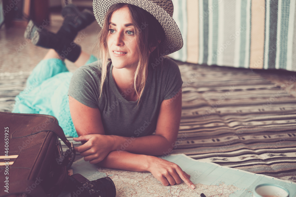 Beautiful young woman lying on carpet and pointing on atlas. Young woman in hat planning for location on map for vacation. Thoughtful woman daydreaming her travel plans while pointing on map