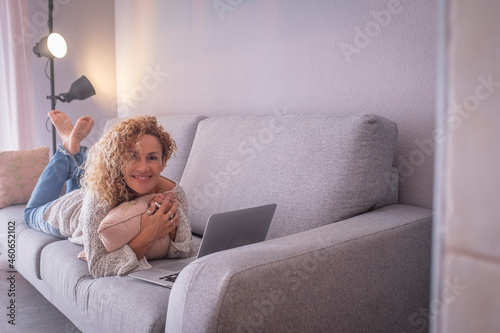 Beautiful young woman working by lying in front of laptop on sofa. Portrait of happy businesswoman working from home. Woman spending leisure time using laptop on sofa in living room