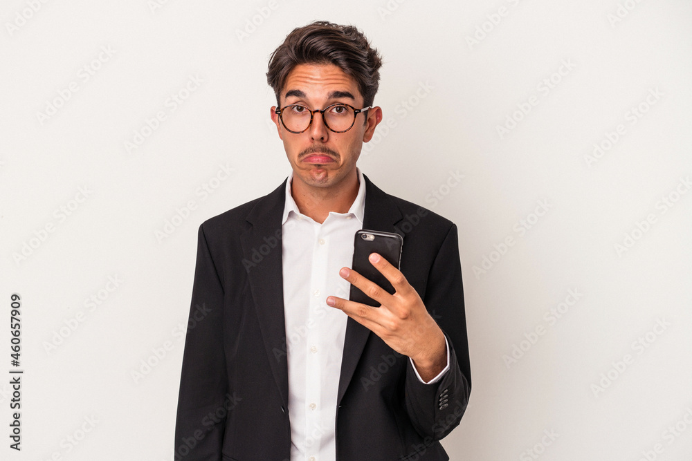Young mixed race business man holding mobile phone isolated on white background shrugs shoulders and open eyes confused. Young mixed race business man holding mobile phone isolated on white background shrugs shoulders and open eyes confused.